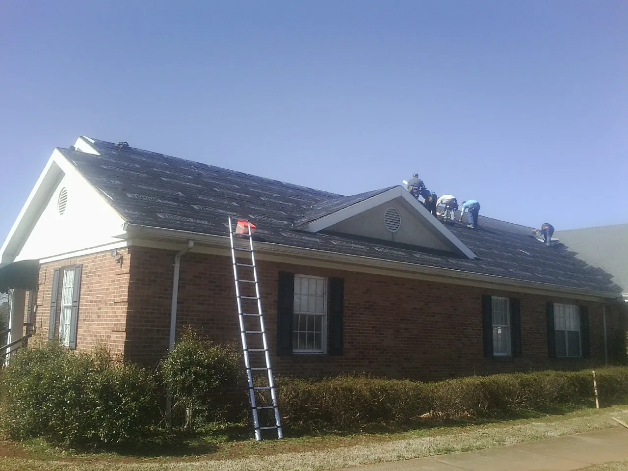 Crew tearing shingles off a brick ranch roof with a ladder set — Metal Roof Repair work by our crew in Satsuma