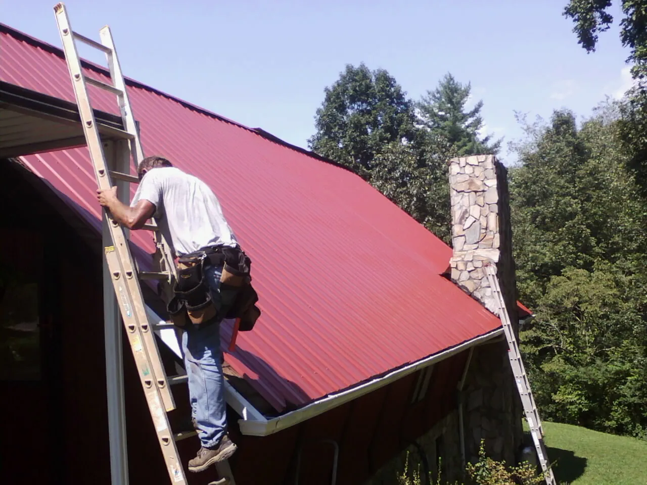 Roofer on a ladder beside a red metal roof and stone chimney — Soffit & Fascia Repair by Roofing Brothers in Satsuma
